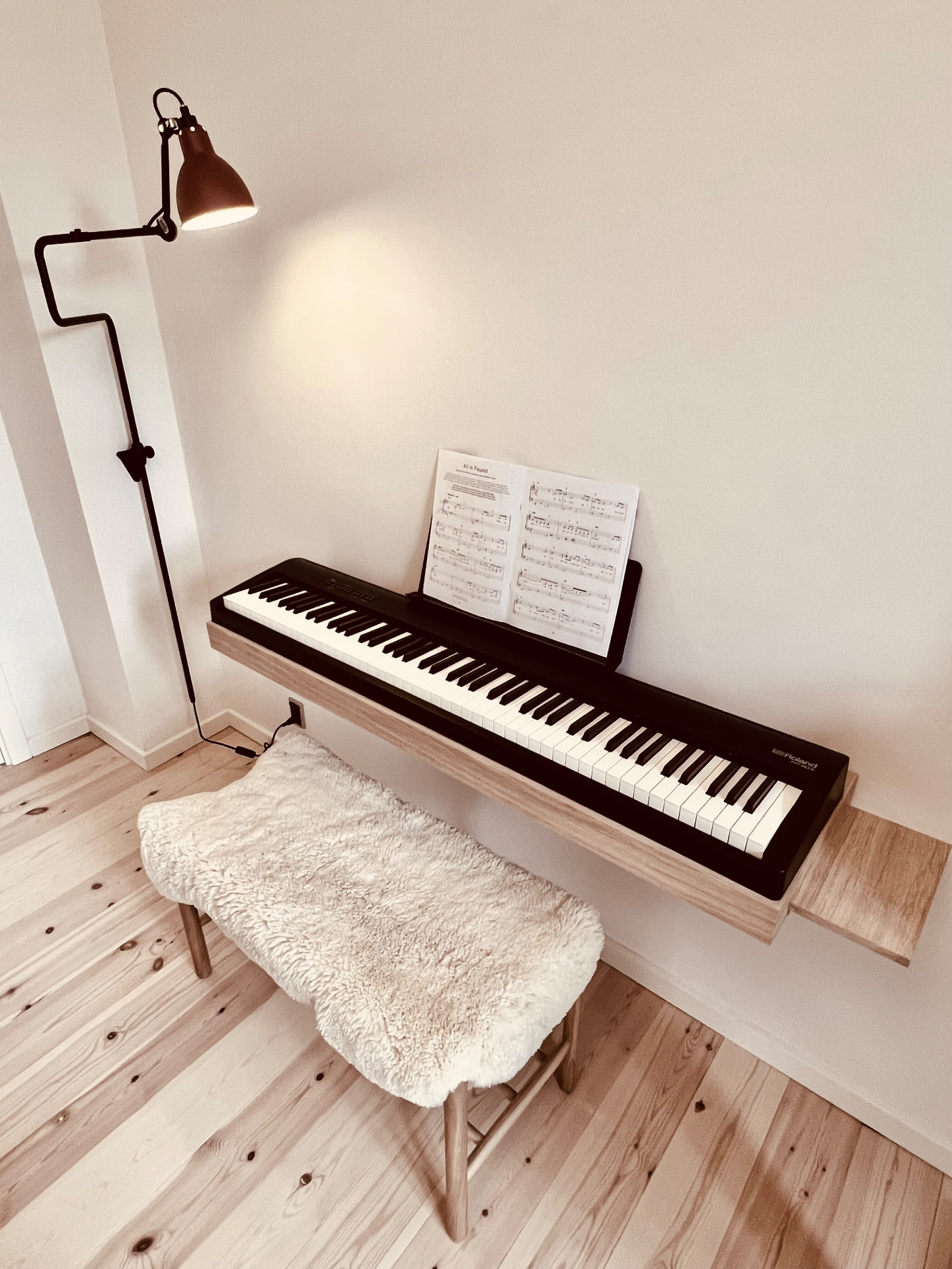 Customer photo of a Roland digital piano on a wall-mounted oak piano stand with side shelf, sheet music and a sheepskin bench